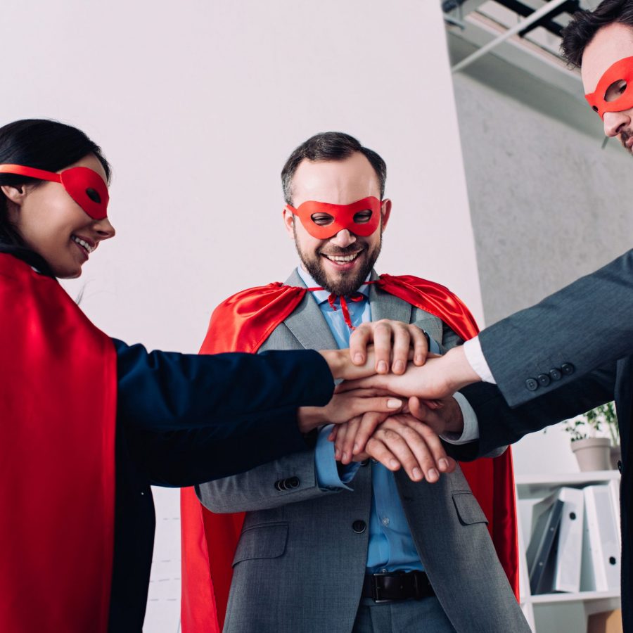 happy super businesspeople in masks and capes putting hands together in office